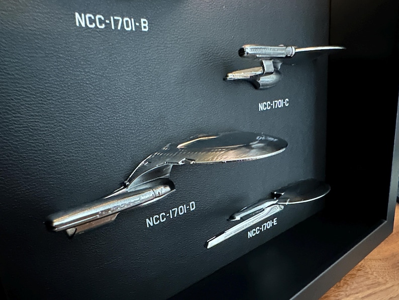 K&ouml;nnte beinhalten: Eine Pr&auml;sentation von vier silberfarbenen Raumschiffmodellen, die jeweils mit "NCC-1701" und einem Buchstaben gekennzeichnet sind. Die Modelle sind auf einer strukturierten schwarzen Oberfl&auml;che montiert, mit wei&szlig;em Text. Die Schiffe befinden sich in einer schwarzen Vitrine.