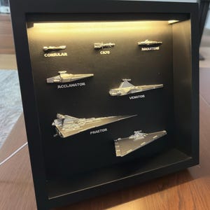 Puede incluir: Una exhibición enmarcada con naves espaciales plateadas en miniatura, incluyendo "Consular", "CR70", "Arquitens", "Acclamator", "Venator", "Praetor", y otra nave. Las naves están montadas sobre un fondo negro dentro de un marco negro.