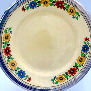 May include: A vintage cream-colored plate with a floral design. The plate features a border of blue, yellow, red, and blue flowers with green leaves. The plate is displayed on a gold-colored stand.