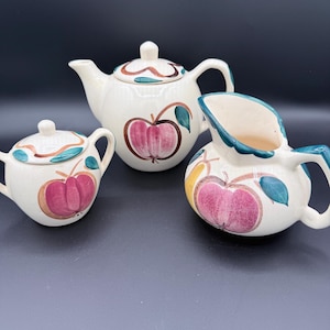 May include: A vintage cream-colored tea set featuring a teapot, sugar bowl, and creamer. Each piece is adorned with a hand-painted apple design in shades of pink, brown, and green. The set is ideal for serving tea or coffee.