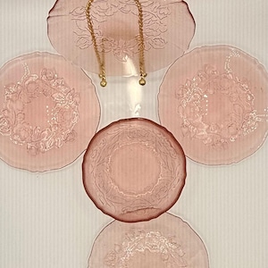 May include: A collection of pink glass plates with floral designs. The set includes various sizes, with a larger, irregularly shaped plate at the top, connected by a gold chain. The plates are arranged on a white surface.