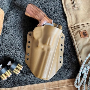 May include: A tan holster with a handgun and a wooden grip. Two black speed loaders with bullets are next to the holster. The items are on a gray, textured surface, with a brown jacket and rope in the background.