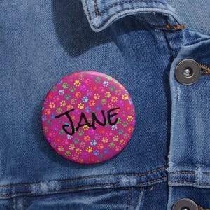 May include: A circular, pink badge featuring a pattern of colourful paw prints. The name "JANE" is written in black script across the centre. The badge is set against a denim jacket.