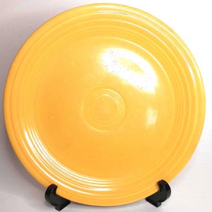 May include: A bright yellow ceramic plate with concentric rings and a central indentation. The plate is displayed on a black stand, showcasing its vibrant color and classic design. This vintage dish is perfect for serving meals.