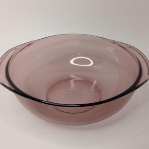 May include: A round, translucent purple glass bowl with integrated side handles. The base of the bowl features embossed text that reads PYREX, MADE IN USA, and 1.5 L. The glass has a smooth, polished finish and a wide, open top design.