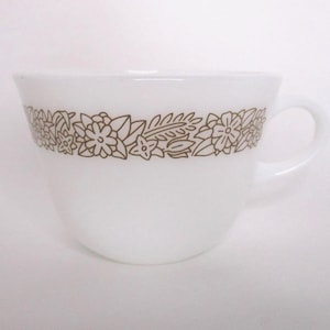 May include: White teacup with a brown floral design around the rim. The cup has a curved handle and a wide opening. The floral pattern includes various flower and leaf shapes. The cup is made of a smooth, opaque material.