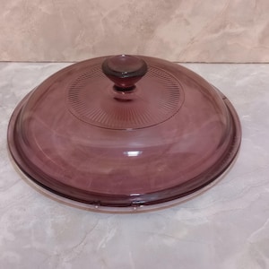 May include: A round, transparent, purple glass lid with a matching knob. The lid has a series of concentric circles and radial lines emanating from the center. The knob is a darker shade of purple.