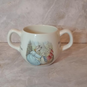 May include: White ceramic baby cup with two handles. The cup features a colorful illustration of rabbits in a garden setting. The illustration includes text. The cup has a rounded shape.