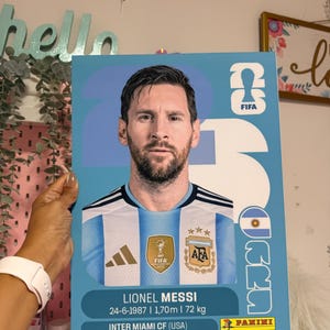 May include: A light blue and white card featuring Lionel Messi, with the FIFA and AFA logos. The card includes Messi's birthdate, height (1.70m), and weight (72 kg). The Panini logo is visible.