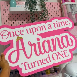 May include: A custom-cut sign with a white background and a thick hot pink border. The text reads "Once upon a time, Ariana Turned ONE" in a mix of script and block fonts. A small gold-colored crown icon sits above the name Ariana.