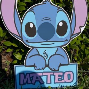 May include: A blue cartoon character yard sign with large ears and the name "MATEO" in pink letters on a blue background. The sign is placed in grass with green foliage in the background.