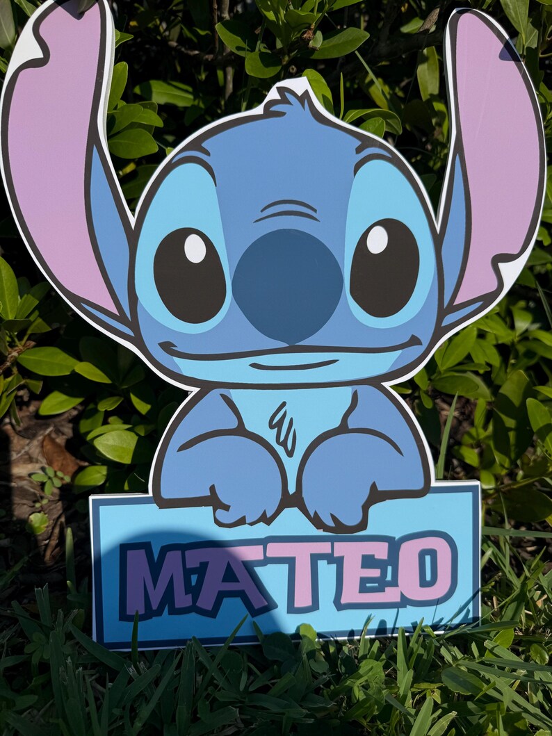 May include: A blue cartoon character yard sign with large ears and the name "MATEO" in pink letters on a blue background. The sign is placed in grass with green foliage in the background.
