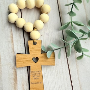Puede incluir: Una cruz de madera con un corazón recortado y la inscripción "Trust in the Lord with all your Heart. Proverbs 3:5." está unida a una cadena de cuentas de madera de color claro. La cruz es de madera marrón claro.