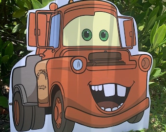 May include: A cartoon cutout of Tow Mater from the movie Cars. The character is orange with a smiling face, large teeth, and green eyes. The cutout is standing in front of green foliage.