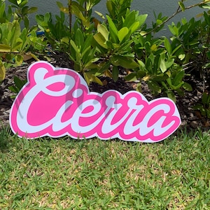 May include: A pink sign with the name "Cierra" in a cursive font with a white outline. The sign is placed on a grassy lawn in front of green foliage.