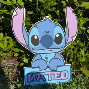 May include: A blue and pink cartoon character cutout with the name "MATEO" in pink letters on a blue sign. The character has large ears, big eyes, and a friendly expression. The cutout is placed in a grassy area with green foliage in the background.