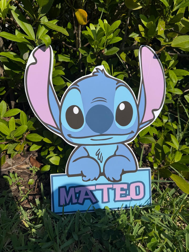 May include: A blue and pink cartoon character cutout with the name "MATEO" in pink letters on a blue sign. The character has large ears, big eyes, and a friendly expression. The cutout is placed in a grassy area with green foliage in the background.