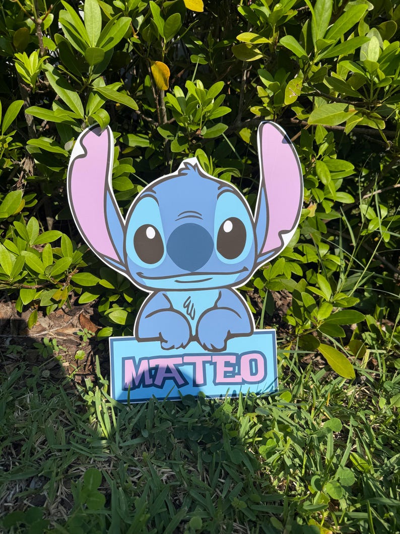 May include: A blue cartoon character cutout with large ears and a sign that reads "MATEO" in pink and blue. The cutout is placed in front of green foliage and grass.