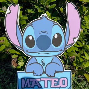 May include: A blue cartoon character with large ears and big eyes, holding a sign that reads "MATEO" in pink letters. The character is set against a backdrop of green foliage and grass.