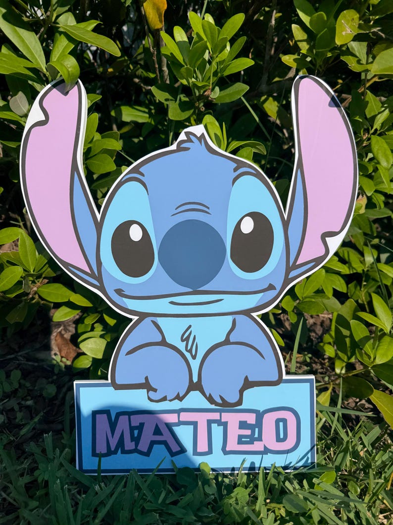 May include: A blue cartoon character with large ears and big eyes, holding a sign that reads "MATEO" in pink letters. The character is set against a backdrop of green foliage and grass.