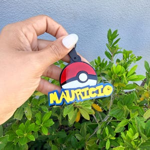 Puede incluir: Un llavero circular con un diseño de bola roja y blanca, borde negro y un círculo blanco central. Debajo de la bola, el nombre MAURICIO aparece en letras mayúsculas amarillas en negrita con un contorno azul sobre una base negra.