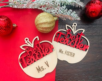 Può includere: Due ornamenti in legno a forma di mela con la scritta "Best Teacher" in rosso. La metà inferiore delle mele è in legno chiaro, con il nome dell'insegnante inciso. Ornamenti natalizi rossi e dorati e verde sullo sfondo.