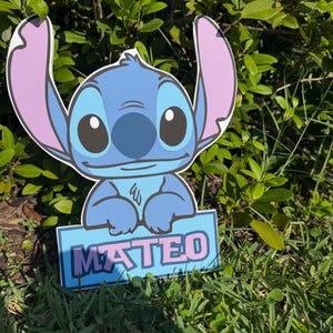 May include: A blue cartoon character cutout with large ears and the name "MATEO" on a sign. The character is set in a grassy area with green foliage in the background. The sign has a blue background with pink lettering.