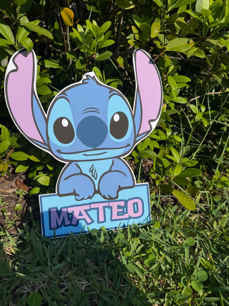 May include: A blue cartoon character cutout with large ears and the name "MATEO" on a sign. The character is set in a grassy area with green foliage in the background. The sign has a blue background with pink lettering.