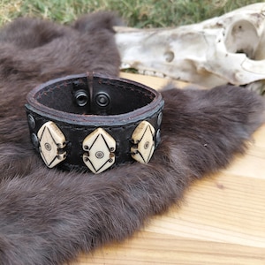 Peut inclure: Un bracelet manchette en cuir noir avec trois ornements crème en forme de bouclier. Chaque bouclier présente un cercle central et un motif géométrique. Le bracelet se ferme avec deux boutons-pression et est présenté sur une surface en fourrure brune.