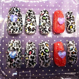 May include: A set of ten artificial nails in a clear plastic container. The nails feature a leopard print design with black, white, and colorful spots. Two nails are red with glitter and a heart.