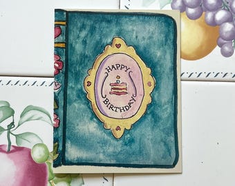 May include: A teal birthday card featuring a gold-framed oval design with a cake illustration and the words "Happy Birthday". The card has a floral and heart design on the left side. The card is on a tiled surface.
