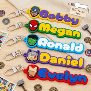 May include: Colorful superhero-themed keychains with names: Bobby, Megan, Ronald, Daniel, and Evelyn. Each keychain features a different character's face and a matching color scheme. Includes metal clasps for easy attachment.