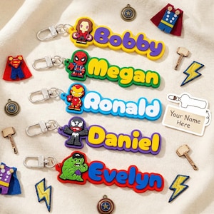Puede incluir: Llaveros con temática de superhéroes con nombres: Bobby, Megan, Ronald, Daniel y Evelyn. Cada llavero presenta un personaje de superhéroe diferente y un nombre en colores vibrantes. Los dijes adicionales incluyen un martillo, un rayo y un escudo.