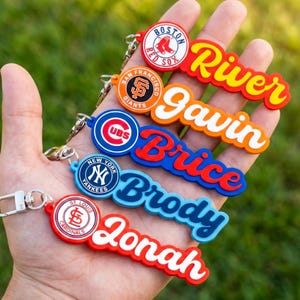 Könnte beinhalten: Eine Sammlung farbenfroher Schlüsselanhänger mit Baseball-Teamlogos und personalisierten Namen. Die Schlüsselanhänger zeigen Namen wie "River", "Gavin", "Brice", "Brody" und "Jonah" in einer verspielten Schriftart, mit Teamlogos wie den Boston Red Sox.