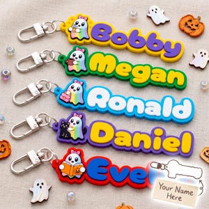 Op de afbeelding: Vijf siliconen sleutelhangers met een cartoongeest met een roze strik en een naam in dikke letters. De namen zijn Bobby, Megan, Ronald, Daniel en Ever. Elke geest houdt een ander voorwerp vast, waaronder een drankje, mok, tas, kat en boek.