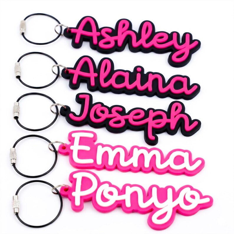 May include: Five personalised name keyrings. Each keyring features a name in a cursive font, with a pink outline and a black base. The names are Ashley, Alaina, Joseph, Emma, and Ponyo. Each keyring has a silver-coloured key ring.