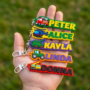 May include: A hand displaying five vibrant keyrings. Each keyring showcases a different vehicle: a fire engine, helicopter, tank, tractor, and a crane. Each keyring has a name in bold letters: Peter, Alice, Kayla, Linda, and Donna.