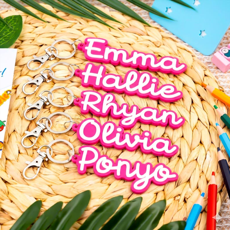 May include: A selection of personalised keyrings, each featuring a name in white script on a pink background. Each keyring has a silver clasp. The names Emma, Hallie, Rhyan, Olivia, and Ponyo are visible. They are arranged on a woven mat.