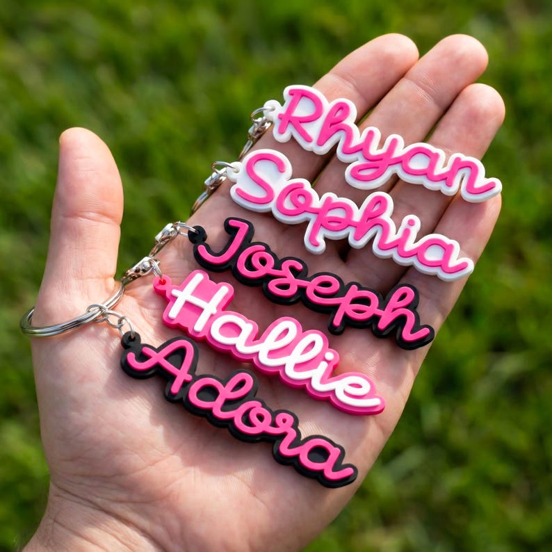 May include: A hand displaying five personalised name keyrings. Each keyring has a different name in a cursive font, with a pink and white colour scheme. The keyrings are attached to silver keyrings.