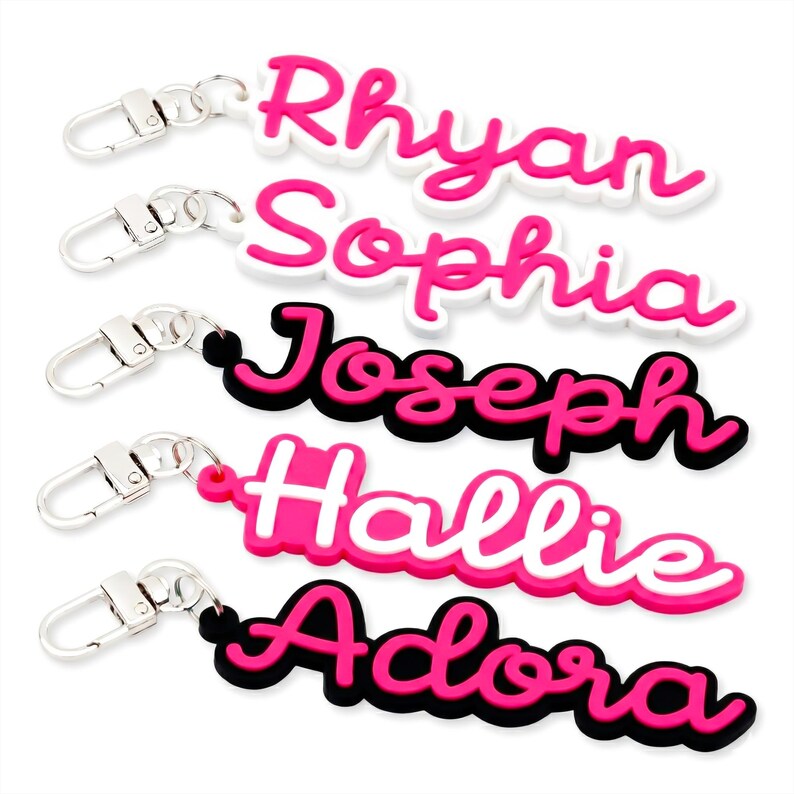 May include: Five personalised name keyrings. Each keyring features a different name in a cursive font: Rhyan, Sophia, Joseph, Hallie, and Adora. The names are in pink with a white or black outline, attached to a silver clasp.