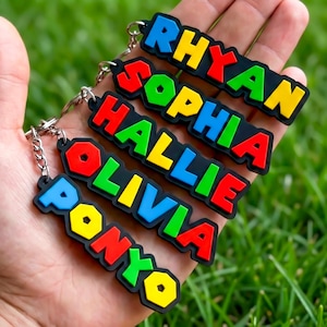 May include: A collection of colorful name keychains. Each keychain features a different name in bold, multi-colored letters against a black background. The names include RHYAN, SOPHIA, HALLIE, OLIVIA, and PONYO. The keychains are held in a hand.