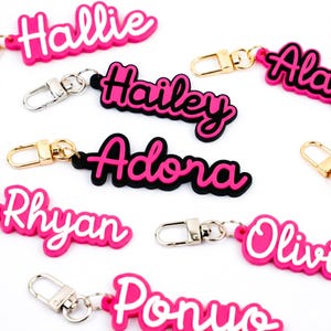 May include: A selection of personalised name keyrings in pink and white. Each keyring displays a different name in a playful, cursive font, outlined in black. The keyrings have silver or gold-coloured clasps.