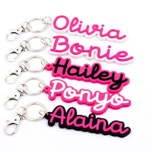 May include: A selection of personalised name keyrings in pink, white, and black. Each keyring displays a different name: Olivia, Bonie, Hailey, Ponyo, and Alaina, in a cursive font. The keyrings have silver-coloured clasps.