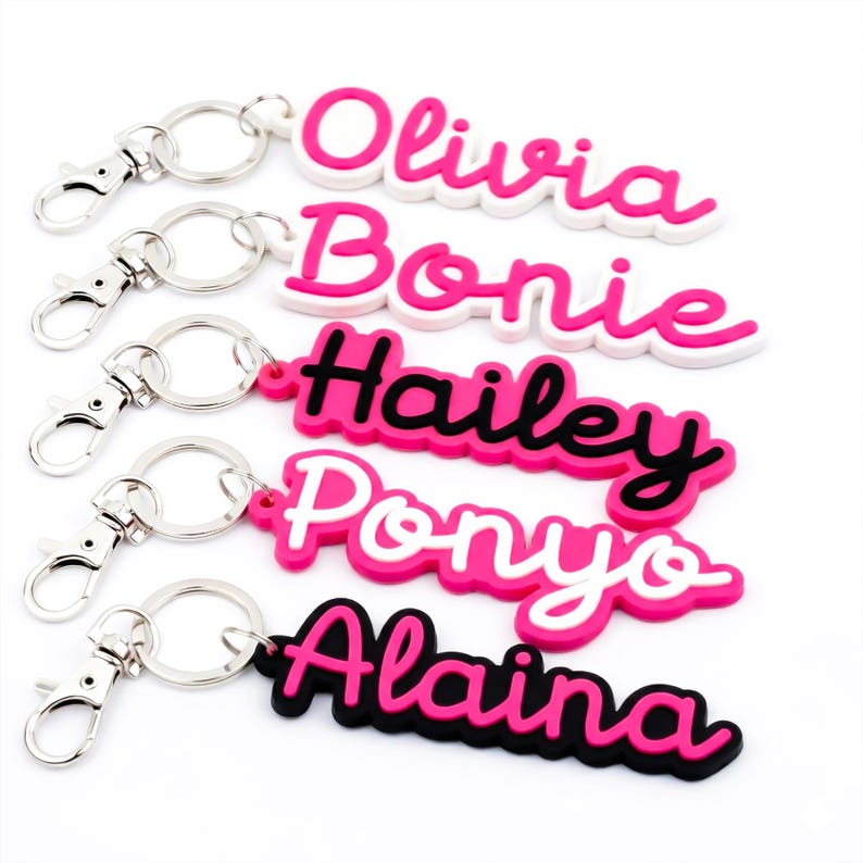 May include: A selection of personalised name keyrings in pink, white, and black. Each keyring displays a different name: Olivia, Bonie, Hailey, Ponyo, and Alaina, in a cursive font. The keyrings have silver-coloured clasps.
