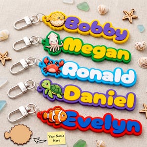Könnte beinhalten: Fünf bunte Silikon-Schlüsselanhänger mit Cartoon-Meerestieren und Namen. Die Anhänger zeigen die Namen Bobby, Megan, Ronald, Daniel und Evelyn in fetten, erhabenen Buchstaben, jeweils befestigt an einem silberfarbenen Metallkarabiner.