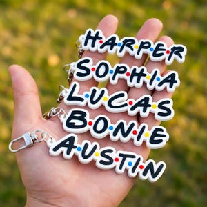 Könnte beinhalten: Weiße Schlüsselanhänger mit Namen in schwarzen Buchstaben, jeweils mit farbigem Punktdesign. Die Namen sind Harper, Sophia, Lucas, Bonie und Austin. Die Schlüsselanhänger werden in einer Hand gehalten.