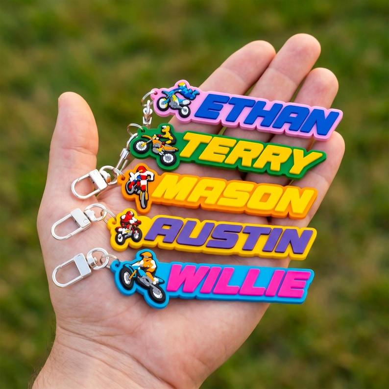 May include: A collection of colorful name keychains featuring a motocross theme. Each keychain has a different name in bold, block letters: Ethan, Terry, Mason, Austin, and Willie. Each keychain has a small motorcycle graphic and a silver clasp.