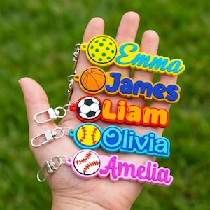 Könnte beinhalten: Eine Sammlung bunter Schlüsselanhänger mit Sportbällen und Namen. Die Schlüsselanhänger zeigen einen Pickleball, Basketball, Fußball, Baseball und Namen wie Emma, James, Liam, Olivia und Amelia. Jeder Schlüsselanhänger hat einen silbernen Verschluss.