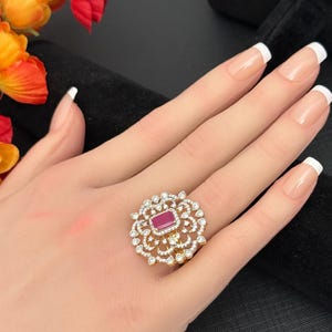 May include: A gold-toned ring with a central rectangular red stone, surrounded by smaller clear stones in a floral design. The ring is displayed on a hand with manicured nails, set against a black background.