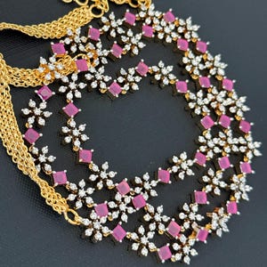 May include: A gold-toned necklace with a delicate chain and a floral design. The necklace features pink square-cut gemstones and clear, round accent stones. The floral design is arranged in a layered pattern, creating a sparkling and elegant look.